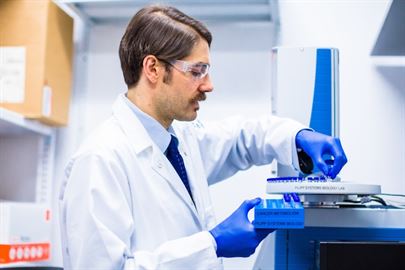 Profile view of man in safety goggles, lab coat, and purple gloves handling tubes in laboratory.