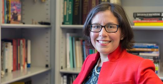 Professor Susana Ramirez in a red jacket and black-rimmed glasses stands in front of bookshelves filled with books.