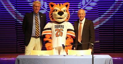 Two men in suits stand beside a tiger mascot wearing a white “TIGERS 26” jersey behind a table on a stage with purple lighting.