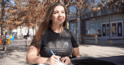 Student stands outdoors on the UC Merced campus holding a notebook and pen, looking off into the distance near a campus walkway lined with leafless trees.