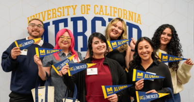 Six UC Merced students pose in front of a University of California, Merced backdrop, smiling and holding blue-and-gold pennants that read “UC Merced Alumni” and “UC Merced.”