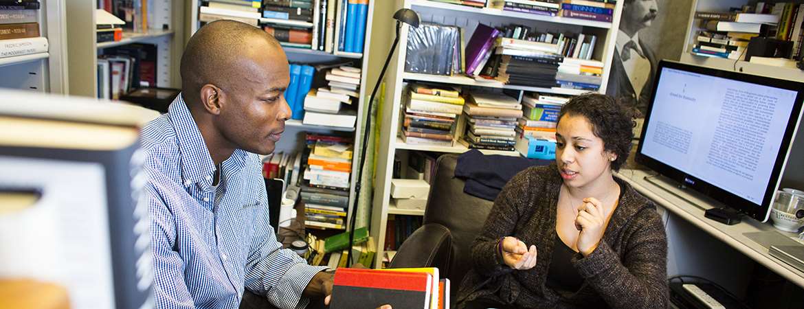 A male professor talks with a female student inside a book-filled office. 