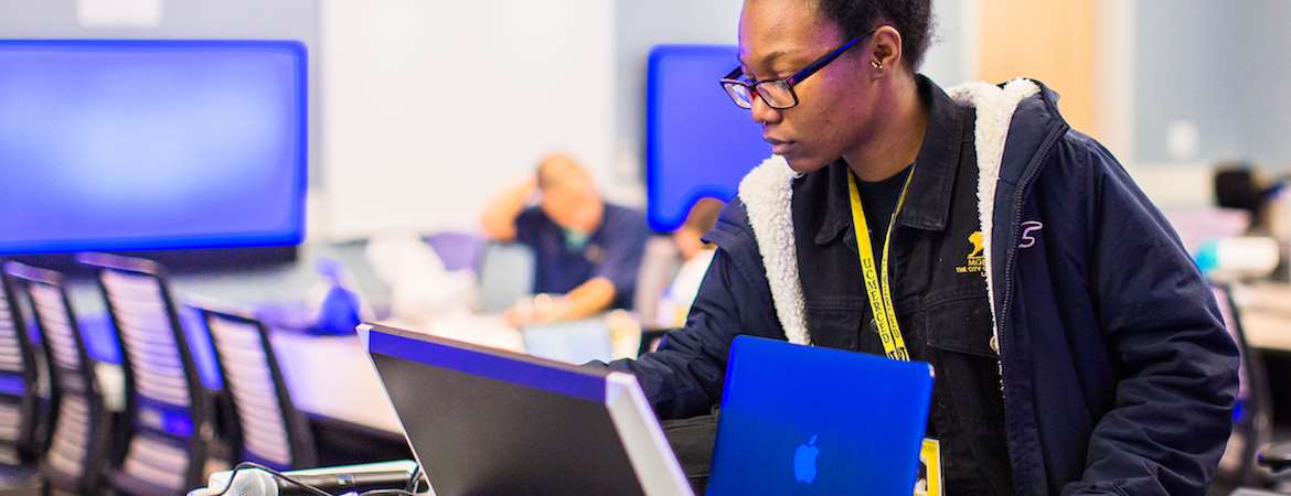 A student works on a blue Apple laptop in the foreground. A conference table with two seated students is behind her. Blue flatscreen TVs line the walls behind the table.