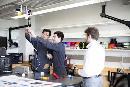 In a laboratory, two men in grey-blue shirts hold a small electronic device at arm's length and inspect it as a man in a white shirt looks on.