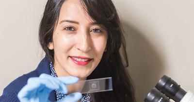 A women in a blue lab coat wearing a blue latex glove holds a microscope slide up to the camera.